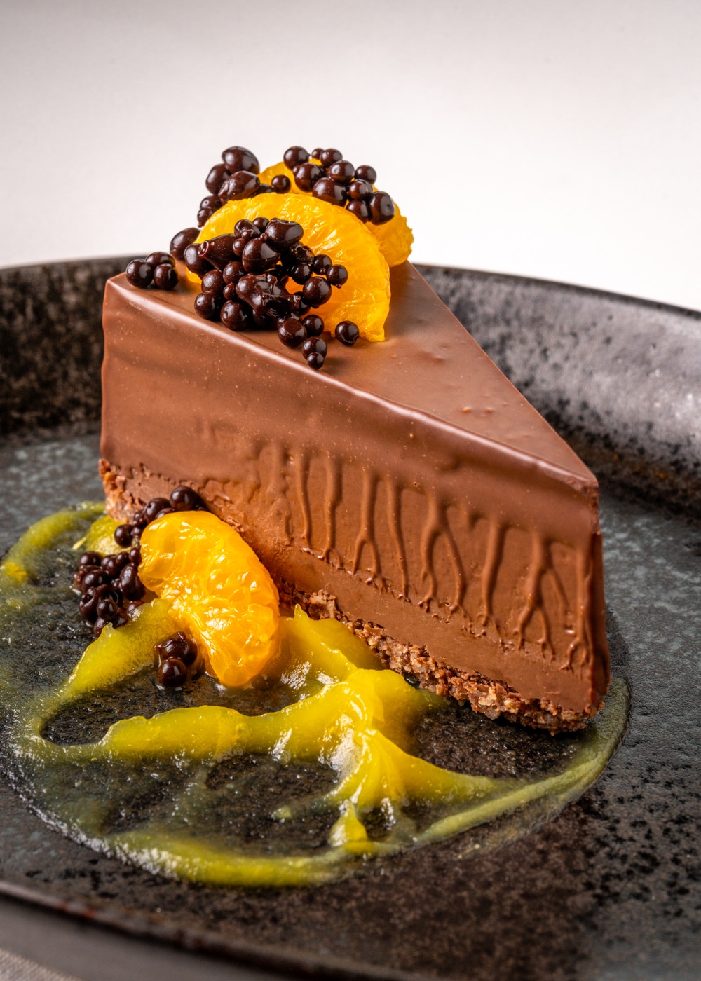 A slice of chocolate cake with a biscuit base, garnished with mandarin orange segments and chocolate pearls, is served on a black plate with a swirl of yellow sauce—a fine dining dessert inspired by twenty-eight Atlantic.