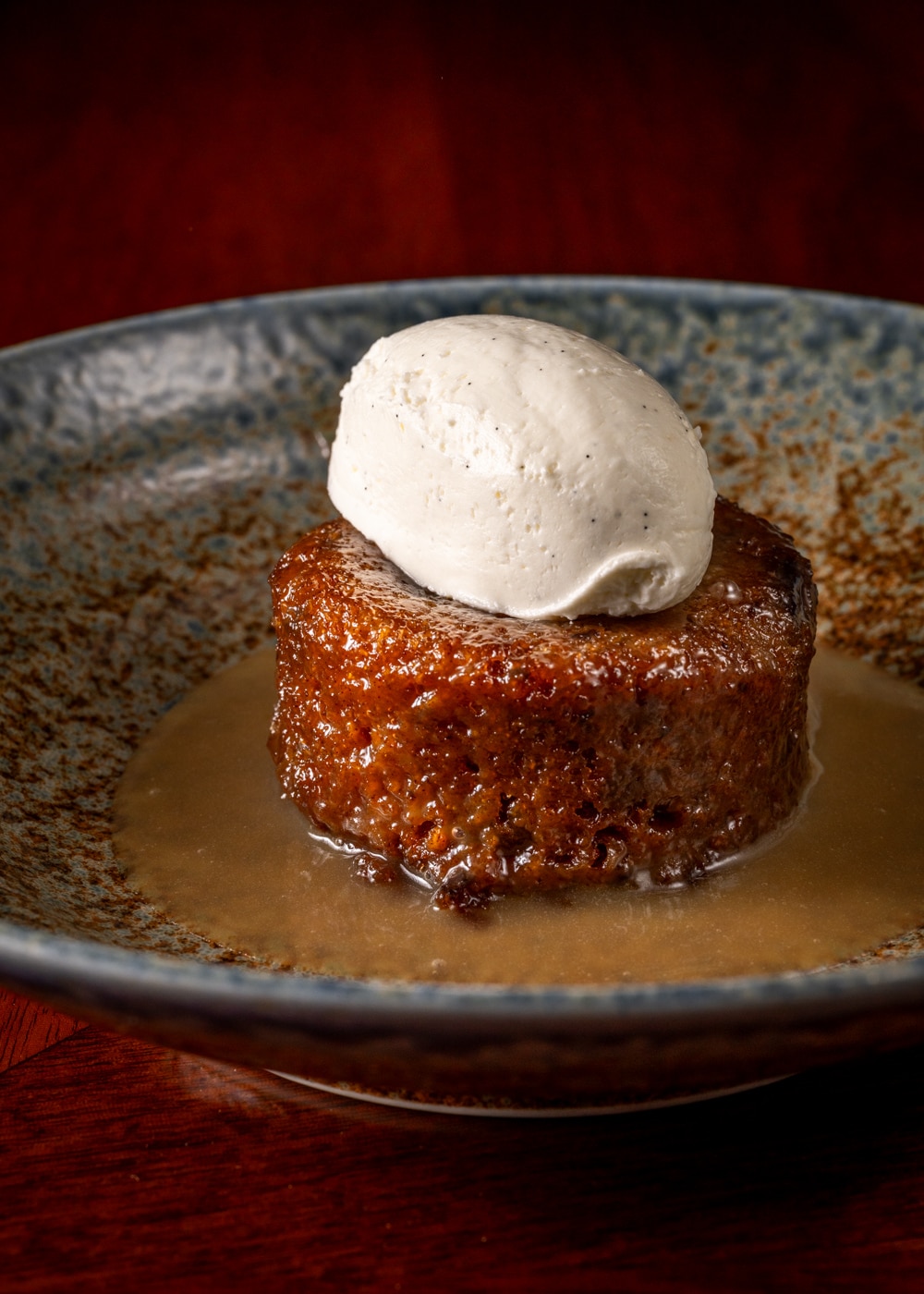A round sticky toffee pudding topped with a scoop of vanilla ice cream, served in a bowl with toffee sauce.