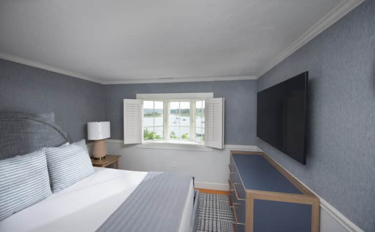 A bedroom with a bed, striped bedding, nightstand with lamp, dresser, large wall-mounted TV, blue walls, and a window with white shutters overlooking water at a charming Cape Cod hotel.