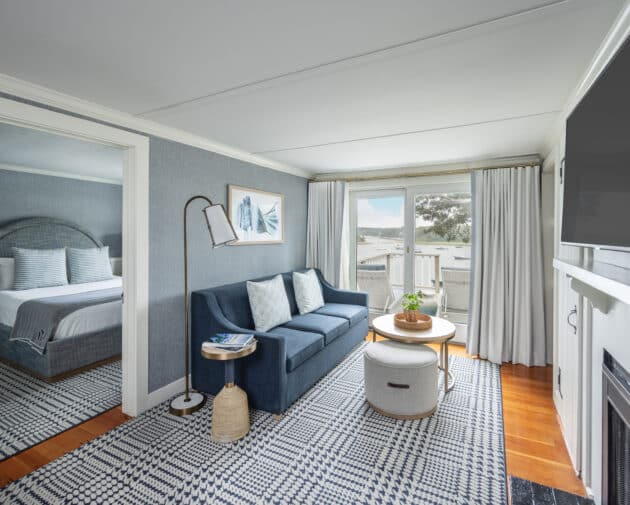 A modern Cape Cod hotel suite with a bedroom and a living area featuring a blue sofa, round coffee table, wall art, and large windows with a view—perfect for relaxing after a day exploring the Cape Cod golf course.