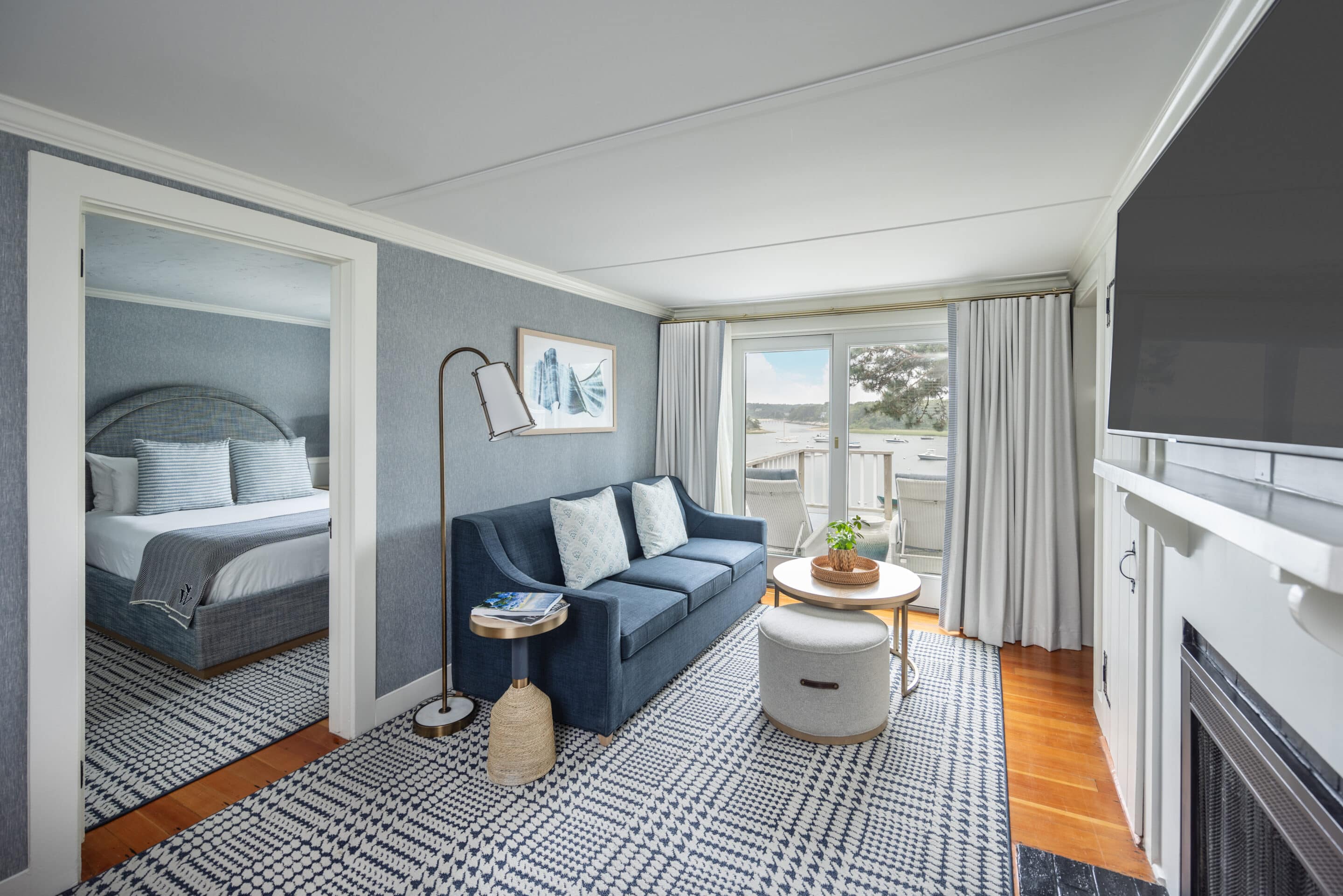 A modern Cape Cod hotel suite with a bedroom and a living area featuring a blue sofa, round coffee table, wall art, and large windows with a view—perfect for relaxing after a day exploring the Cape Cod golf course.