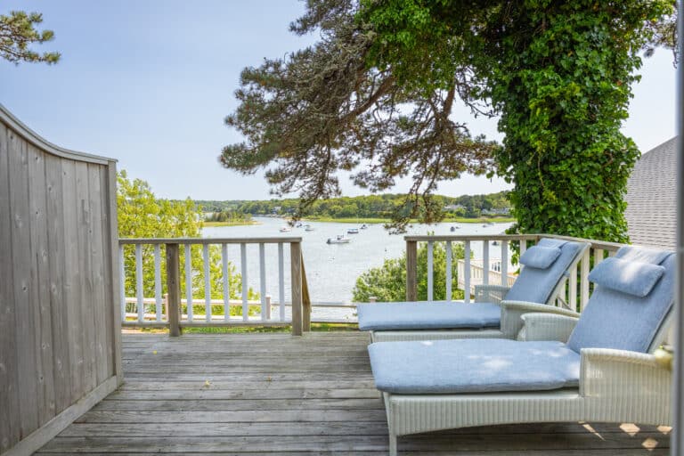 Two cushioned lounge chairs on a wooden deck at 28 Atlantic overlook a calm river with boats, surrounded by trees and greenery on a clear day—an inviting Cape Cod resort setting.