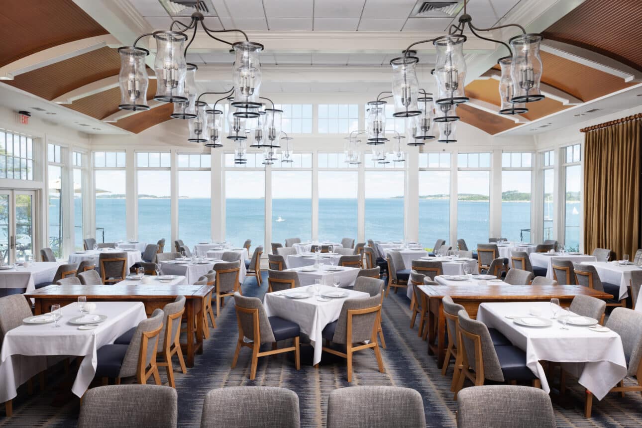 Bright Nantucket restaurant dining room with large windows overlooking a scenic waterfront view. Tables are set with white tablecloths and modern pendant lights, offering a fine dining Cape Cod experience reminiscent of twenty-eight Atlantic.
