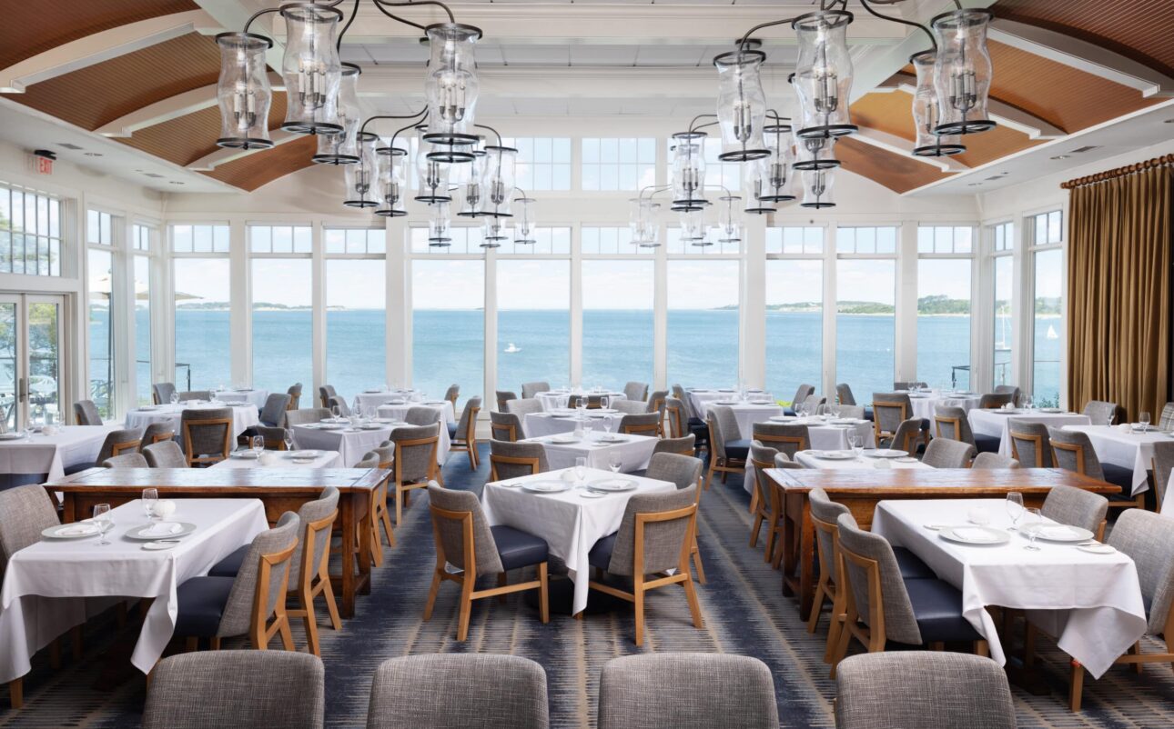Bright Nantucket restaurant dining room with large windows overlooking a scenic waterfront view. Tables are set with white tablecloths and modern pendant lights, offering a fine dining Cape Cod experience reminiscent of twenty-eight Atlantic.