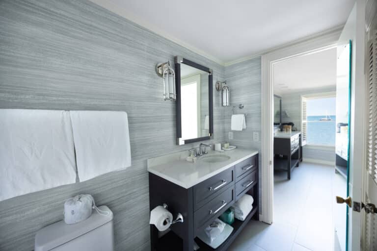 Modern bathroom with double sink vanity, large mirror, towel rack, and an open door revealing a stunning ocean view and a sailboat—perfect for relaxing at a Cape Cod resort like 28 Atlantic.