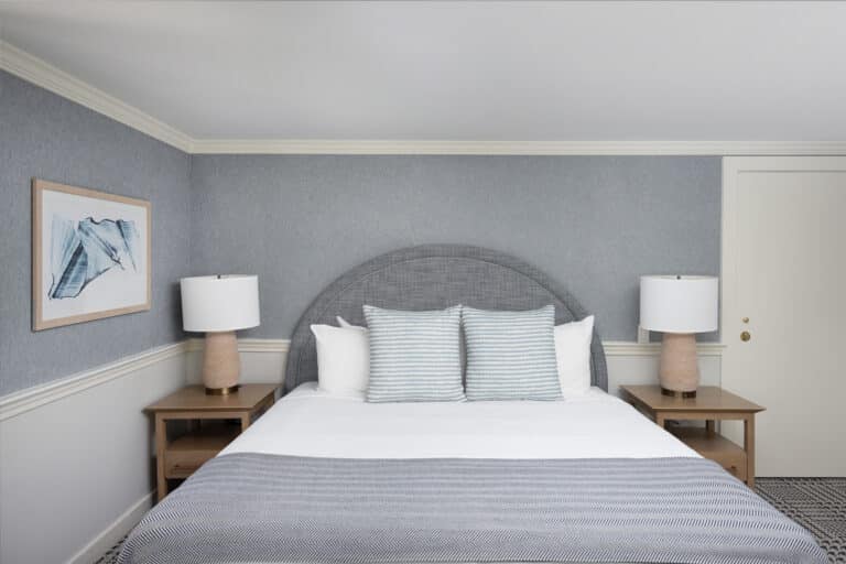 A neatly made bed with three striped pillows, two bedside tables with lamps, a framed abstract art piece, and light gray walls create a serene, minimalist bedroom reminiscent of a Cape Cod resort.