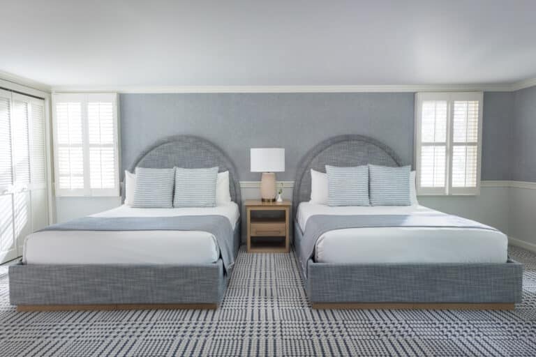A hotel room with two double beds, gray upholstered headboards, a central nightstand with a lamp, striped bedding, and sunlight coming through shuttered windows at the Wequassett Resort and Golf Club.