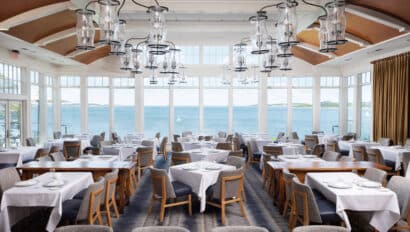Spacious restaurant dining area with white tablecloths, gray chairs, large windows with a view of a body of water, and modern glass chandeliers hanging from the ceiling.