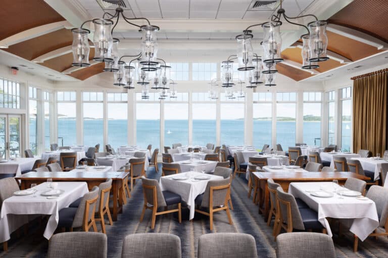 Spacious restaurant dining area with white tablecloths, gray chairs, large windows with a view of a body of water, and modern glass chandeliers hanging from the ceiling.