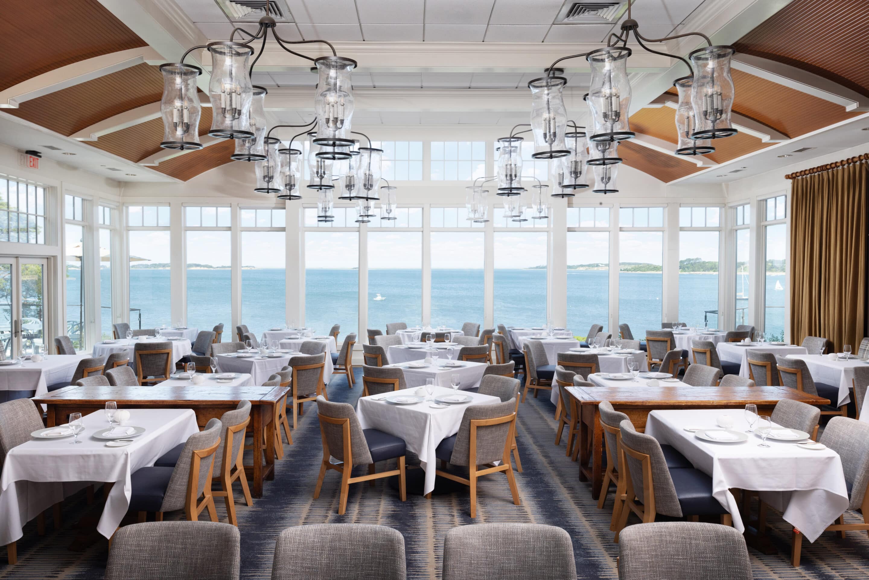 Spacious restaurant dining area with white tablecloths, gray chairs, large windows with a view of a body of water, and modern glass chandeliers hanging from the ceiling.