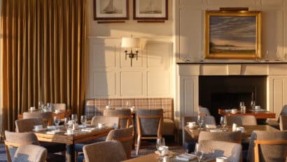 Sunlit dining room at twenty-eight Atlantic with neatly set tables, upholstered chairs, framed sailboat pictures, gold curtains, and a large landscape painting above a black fireplace creates classic Cape Cod restaurant charm.