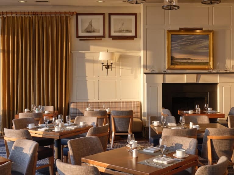 Sunlit dining room at twenty-eight Atlantic with neatly set tables, upholstered chairs, framed sailboat pictures, gold curtains, and a large landscape painting above a black fireplace creates classic Cape Cod restaurant charm.