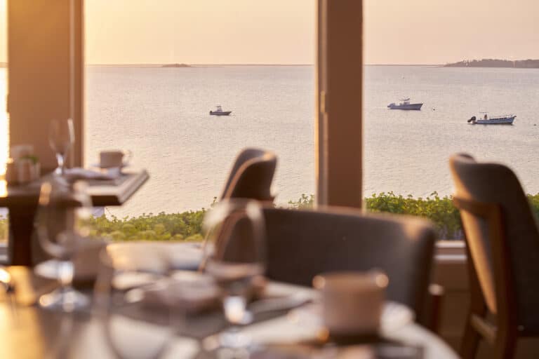 A sunlit restaurant with set tables overlooks a calm body of water with three boats in the distance, seen through large windows.