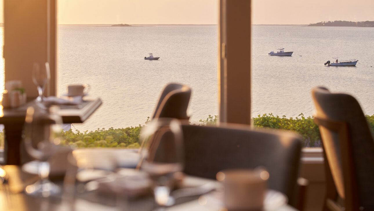 A restaurant dining area overlooks a calm body of water with three boats floating in the distance, viewed through large windows at sunset.