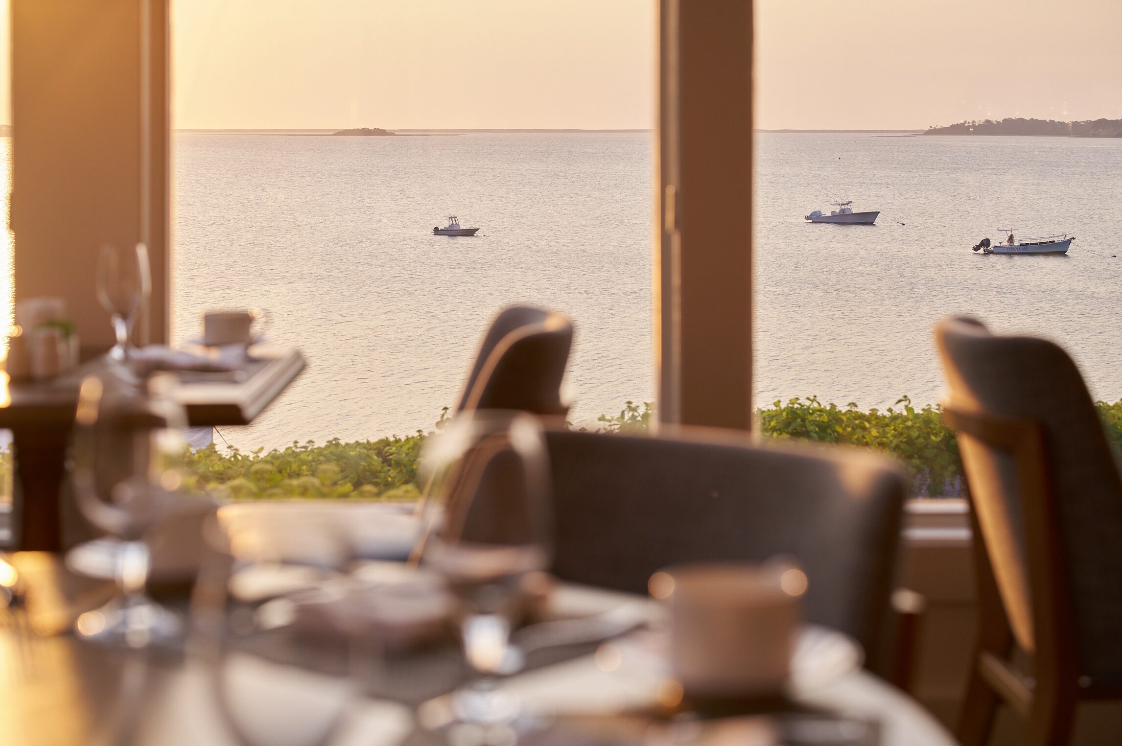 A restaurant dining area overlooks a calm body of water with three boats floating in the distance, viewed through large windows at sunset.