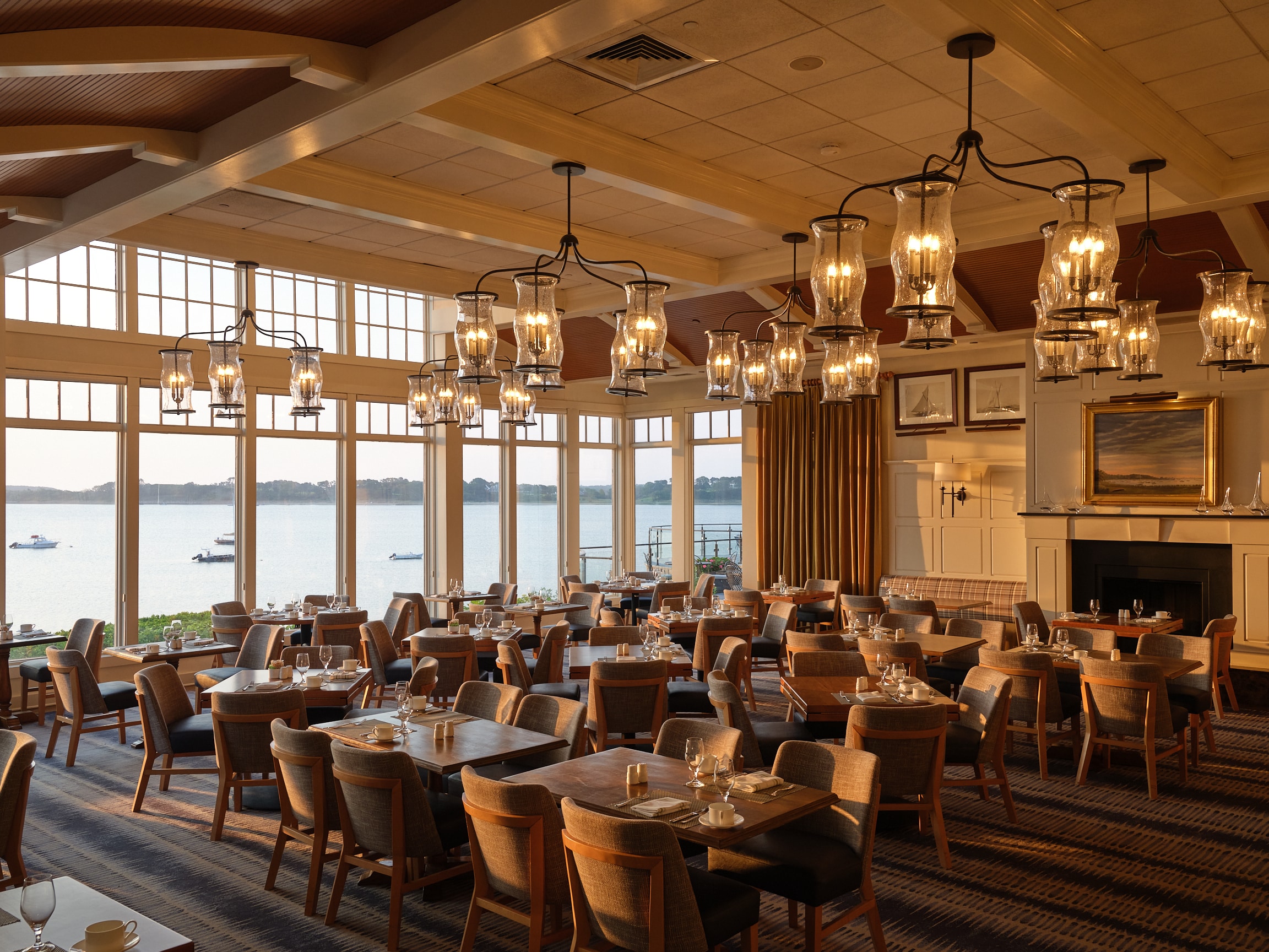 Spacious restaurant dining area with wooden tables and chairs, large windows overlooking a waterfront, and multiple chandeliers providing warm lighting.