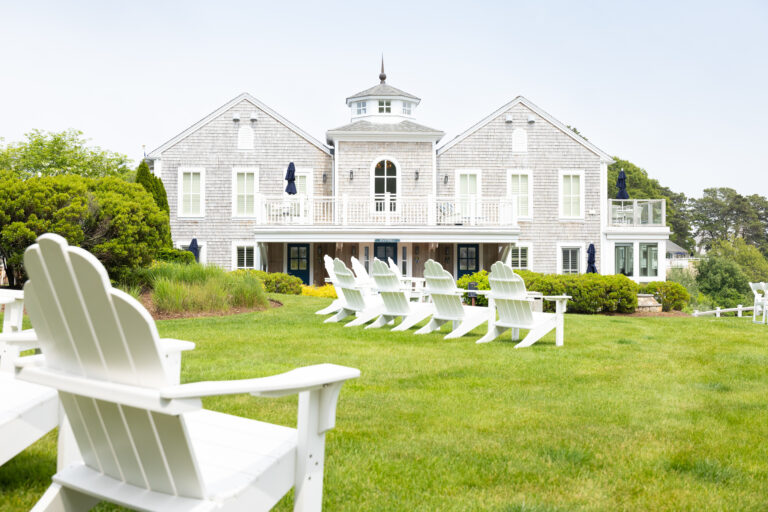 Large gray shingle-style house with white trim, surrounded by a green lawn at Wequassett Resort, with white Adirondack chairs arranged in rows facing the building.