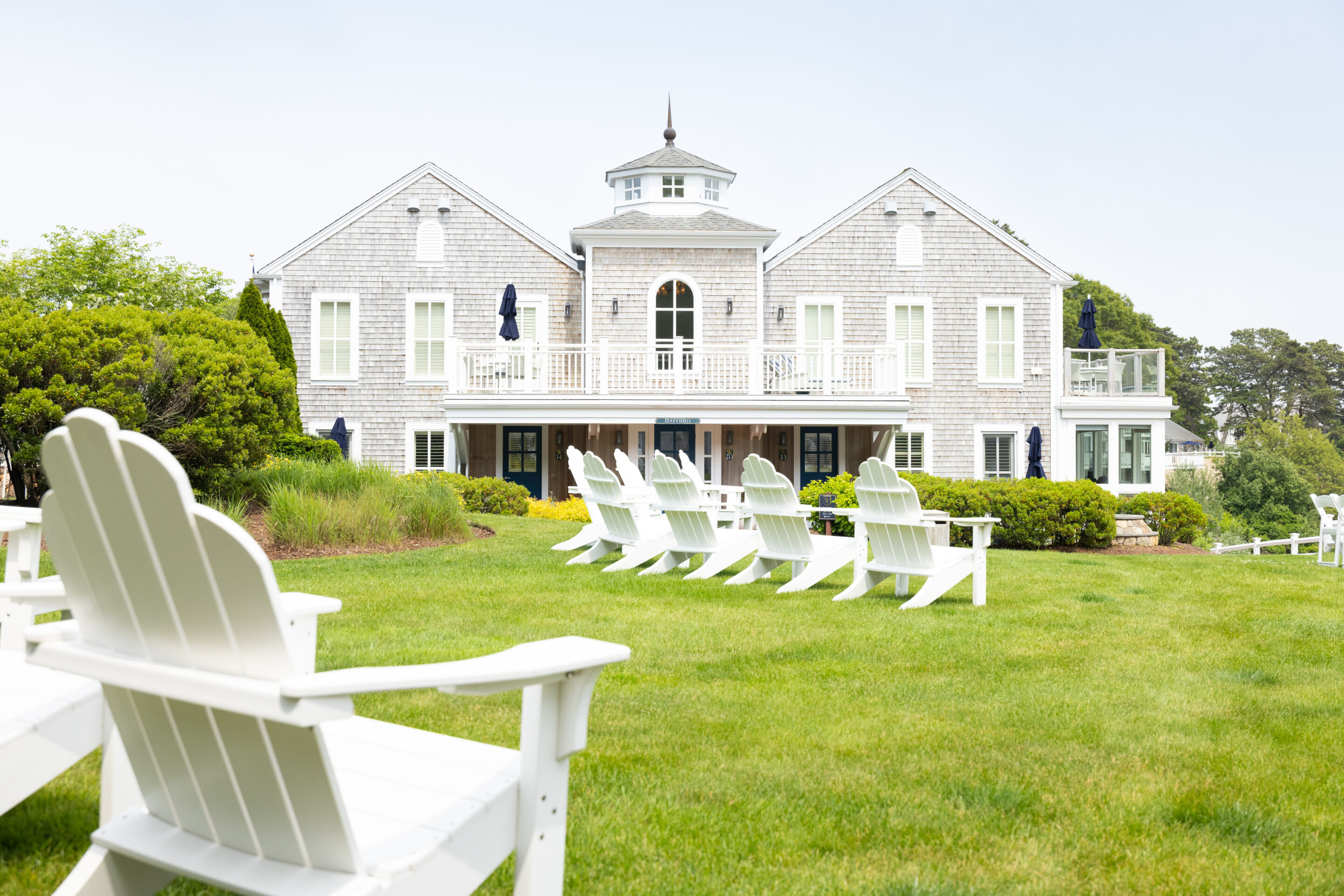Large gray shingle-style house with white trim, surrounded by a green lawn at Wequassett Resort, with white Adirondack chairs arranged in rows facing the building.