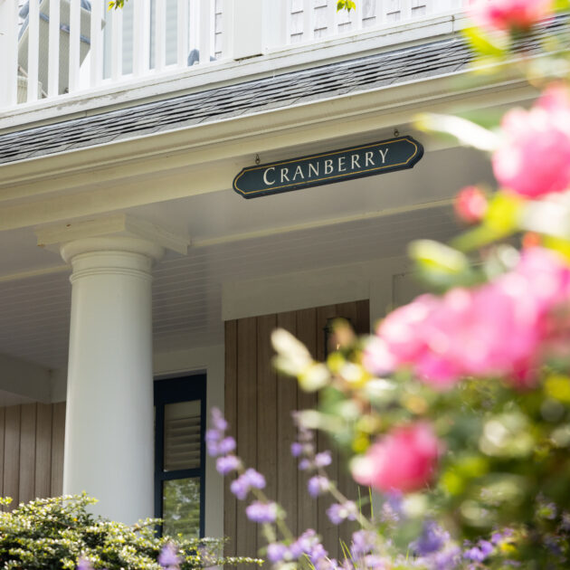 A house porch with a sign labeled "Cranberry" above a column, surrounded by blooming pink and purple flowers.
