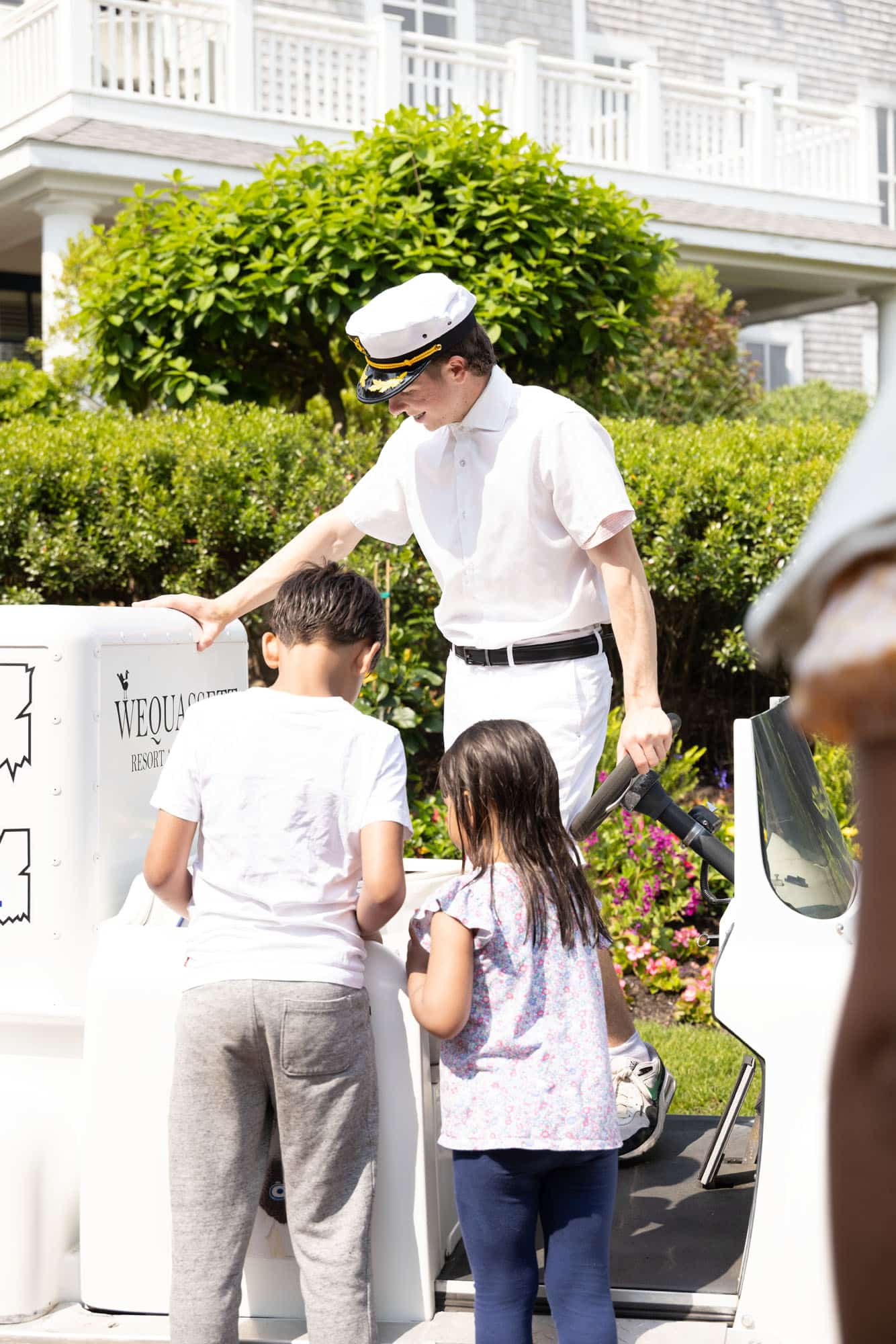 A uniformed attendant helps two children at an outdoor kiosk on a sunny day, with greenery and a building in the background.