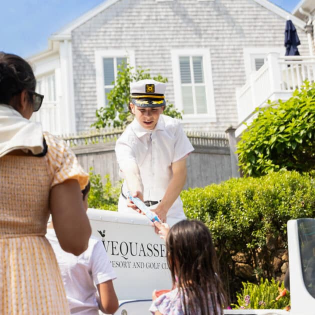 A person in a captain’s hat hands an item to a child near a golf cart outside a gray shingled building. Two other people stand nearby.