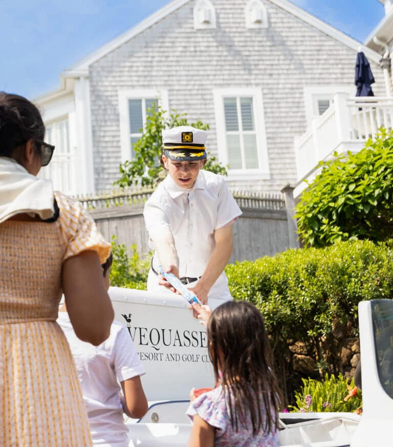 A person in a captain’s hat hands an item to a child near a golf cart outside a gray shingled building. Two other people stand nearby.