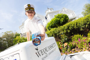A person in a captain’s hat and white uniform hands out an ice cream cup from a cart labeled “Wequassett Resort and Golf Club,” set amid landscaped gardens, where luxurious accommodations await guests.