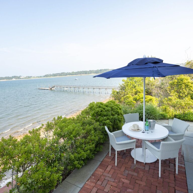 A round white patio table with four chairs and a blue umbrella sits on a brick patio at a Cape Cod hotel, overlooking a calm body of water and long dock. Bushes and trees surround this serene spot near 28 Atlantic.