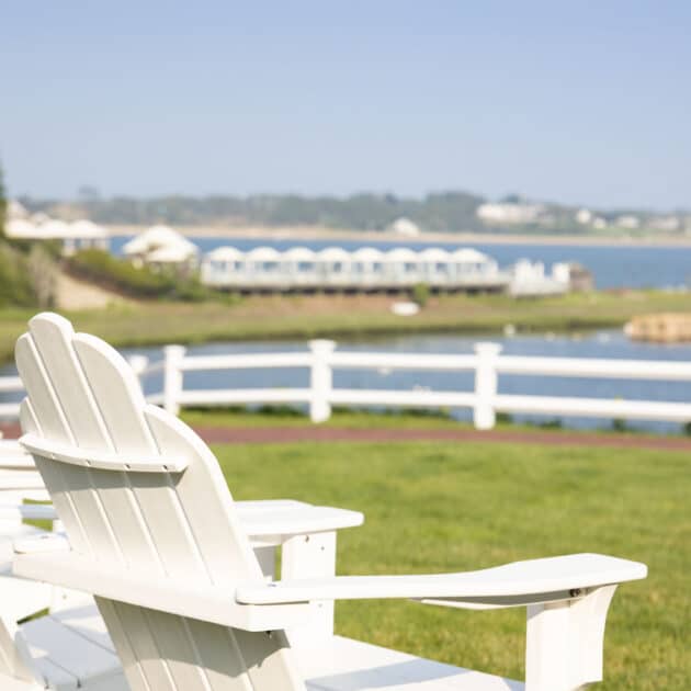 Two white Adirondack chairs sit on a grassy lawn, facing a fence and a body of water with buildings visible in the distance under a clear sky.