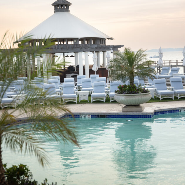 Poolside area with white lounge chairs, a gazebo with a flag on top, and potted plants, set against a calm sky at sunset.