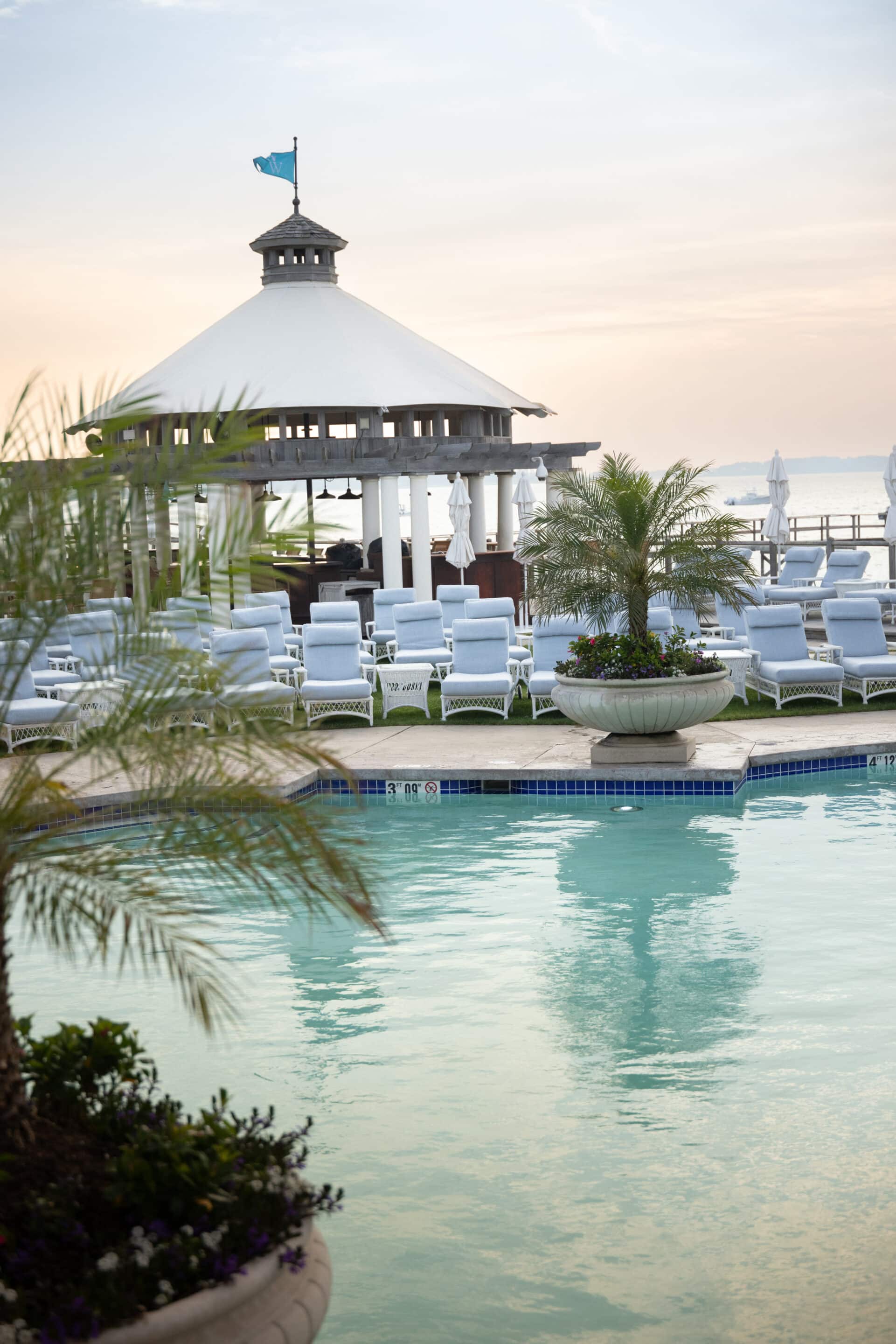 Poolside area with white lounge chairs, a gazebo with a flag on top, and potted plants, set against a calm sky at sunset.