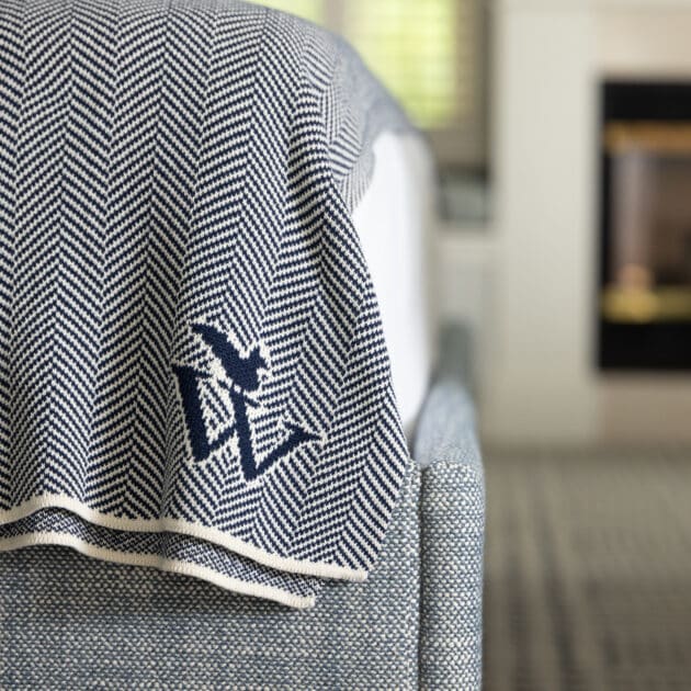 A close-up of a bed with a blue and white herringbone-patterned blanket featuring a bird and initials, next to a lit fireplace in a modern Cape Cod hotel room.
