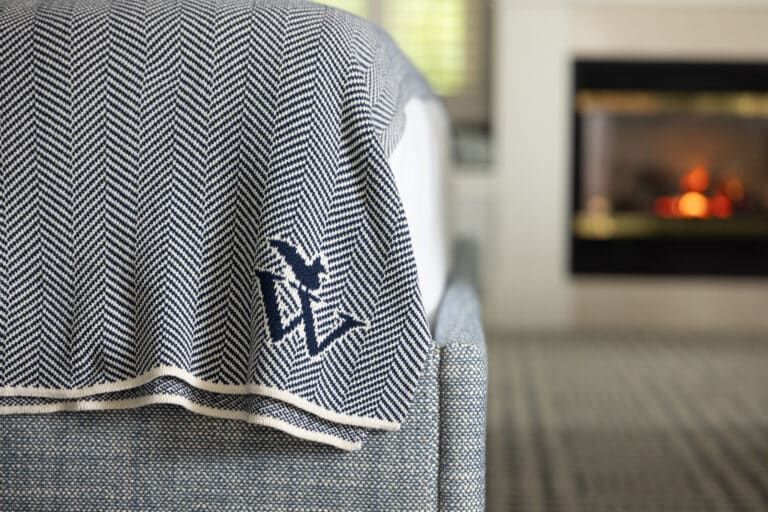 A close-up of a bed with a blue and white herringbone-patterned blanket featuring a bird and initials, next to a lit fireplace in a modern Cape Cod hotel room.