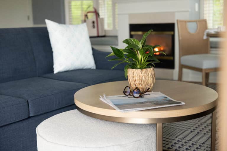 A potted plant, sunglasses, and a newspaper rest on a round coffee table in front of a blue sofa at this inviting Cape Cod resort, with a fireplace and chair adding charm to the cozy setting.