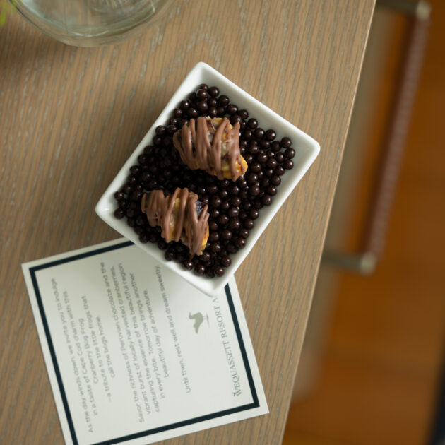 A small square dish with two pieces of chocolate-coated treats on a bed of chocolate pearls sits on a wooden table next to a card and a vase of white flowers.