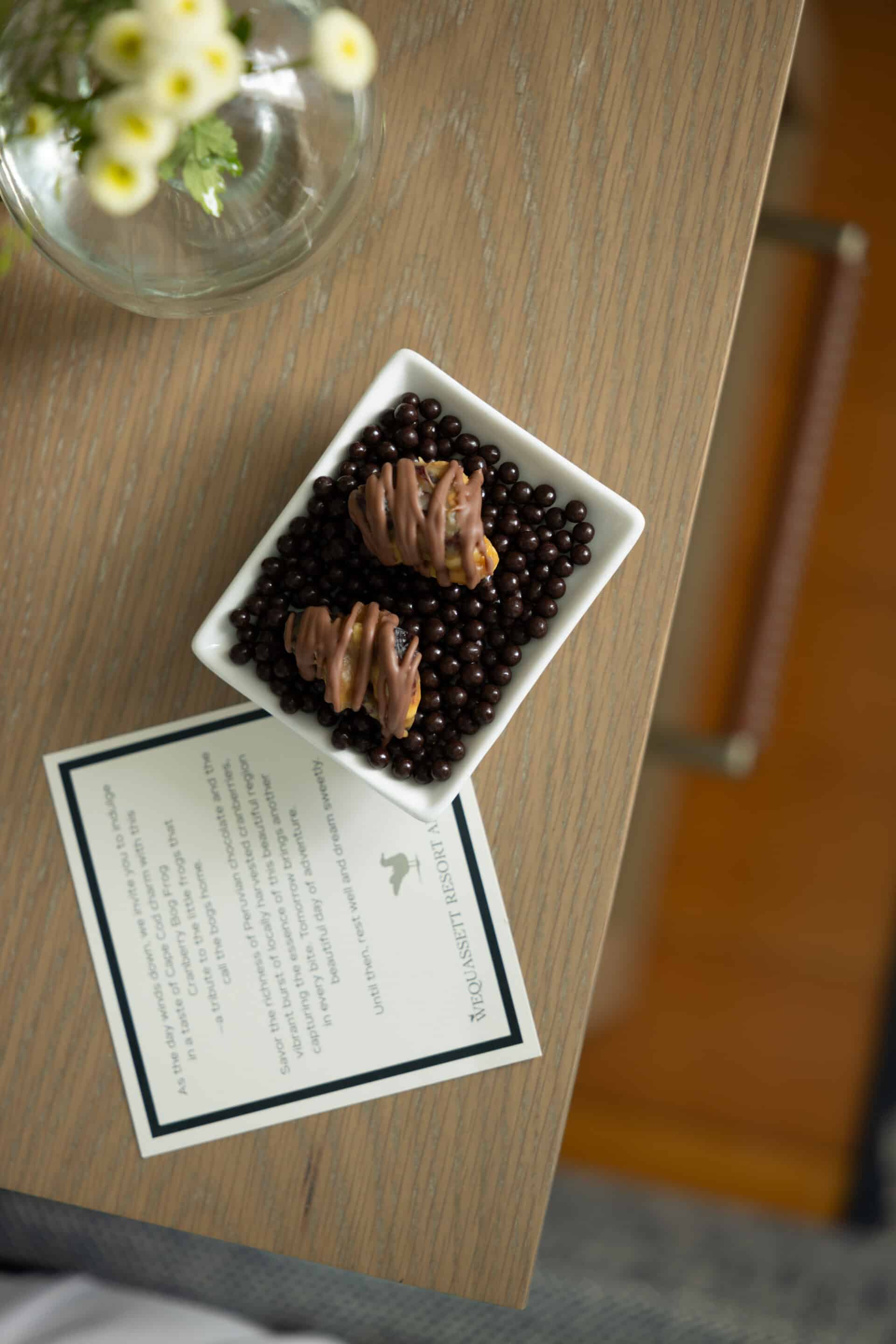A small square dish with two pieces of chocolate-coated treats on a bed of chocolate pearls sits on a wooden table next to a card and a vase of white flowers.