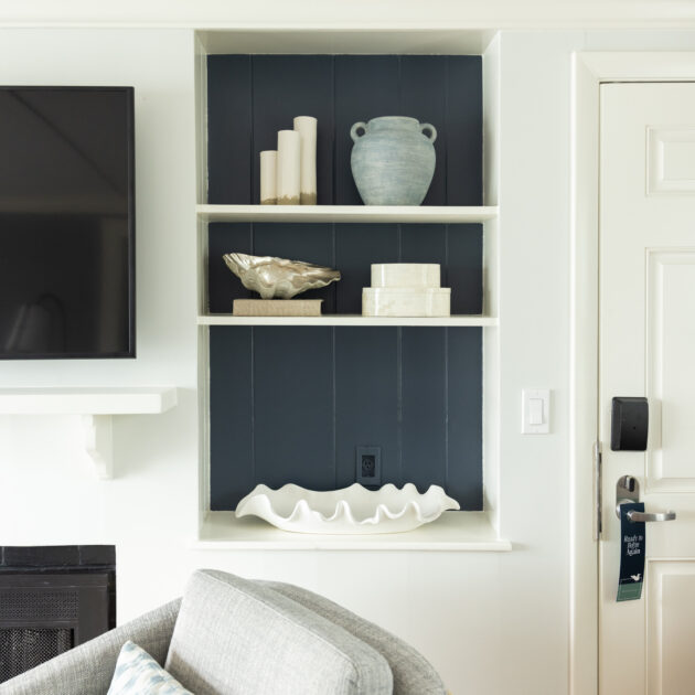Built-in wall shelf with three levels holding decorative objects, including candles, a vase, a bowl, boxes, and a large white shell, next to a TV and a door.