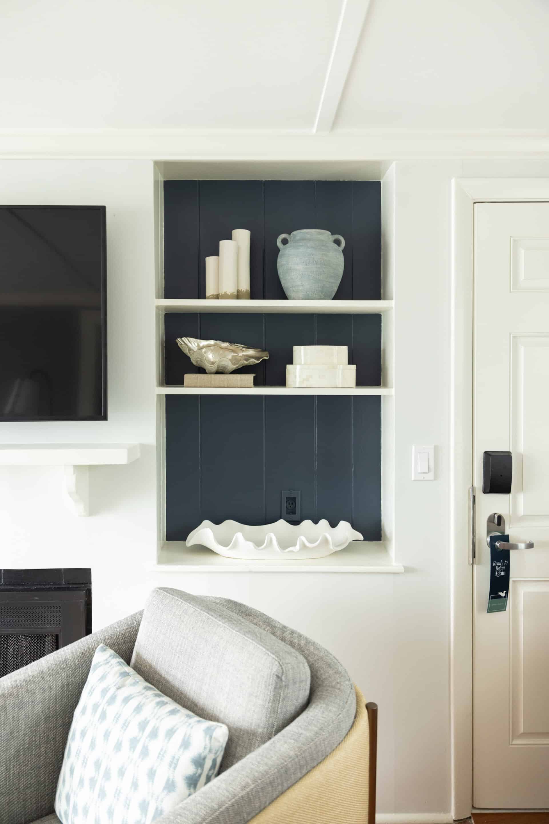 Built-in wall shelf with three levels holding decorative objects, including candles, a vase, a bowl, boxes, and a large white shell, next to a TV and a door.
