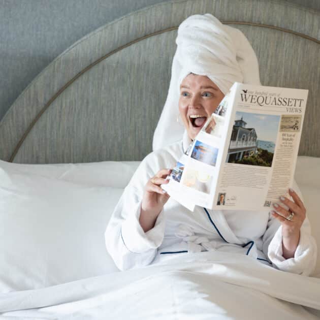 Person in a white bathrobe and towel turban sits in bed, smiling and reading a copy of Wequassett Views magazine.