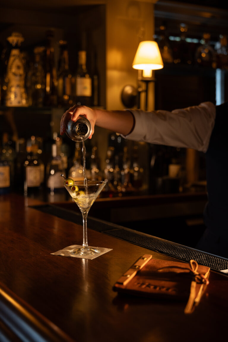 A bartender pours a clear liquid into a martini glass garnished with olives at a dimly lit restaurant Cape Cod bar, with bottles visible in the background.