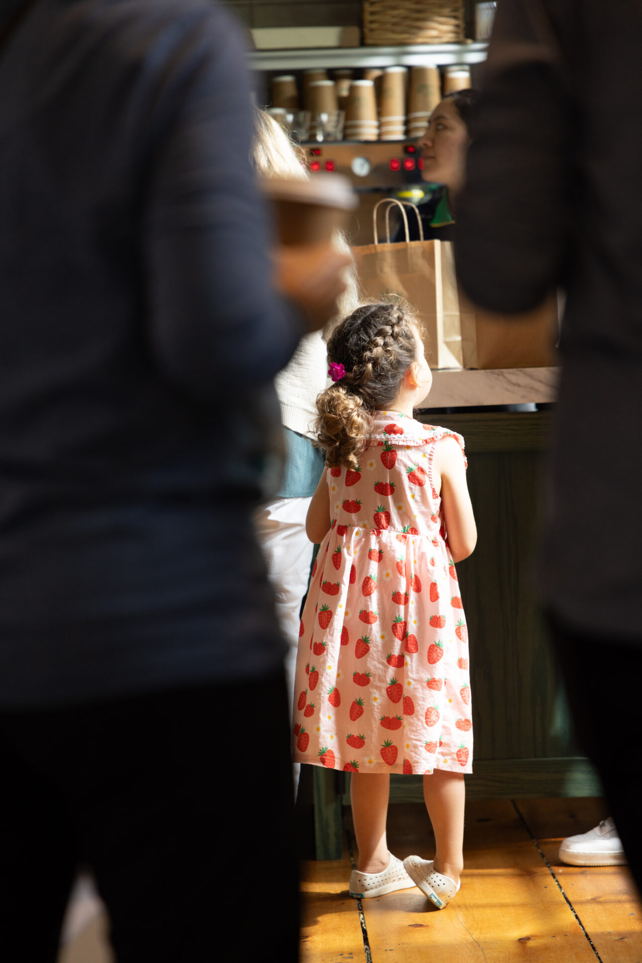 A young girl in a strawberry-print dress stands at a counter, surrounded by adults, in what appears to be a café or restaurant Cape Cod visitors might love.
