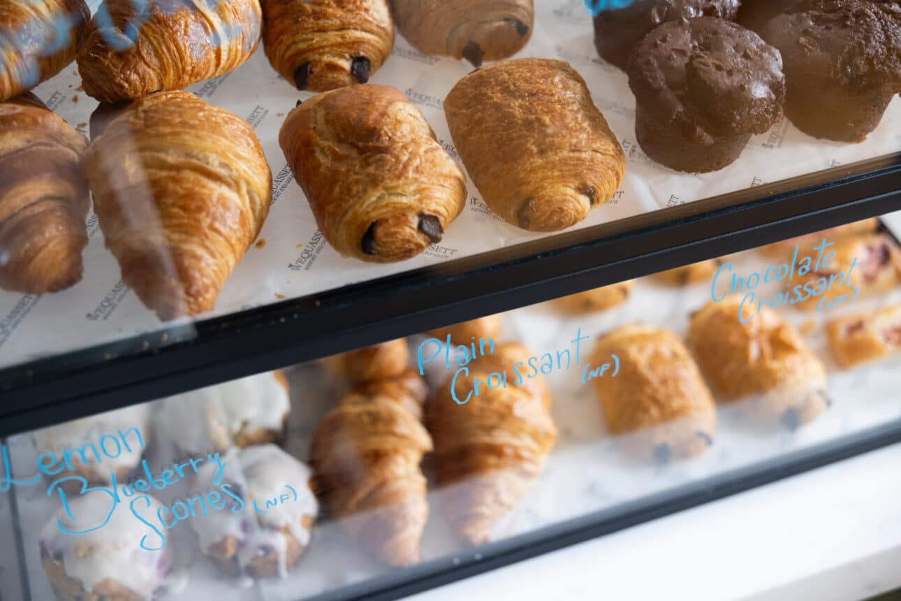 A bakery display case at a restaurant Cape Cod showcases plain croissants, chocolate croissants, chocolate muffins, and lemon blueberry scones behind sparkling glass.