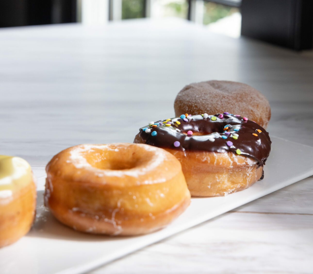 A row of assorted donuts sits on a white rectangular plate, including glazed, chocolate with sprinkles, and cinnamon sugar varieties—perfect for a sweet morning at Wequassett Resort and Golf Club on Cape Cod.