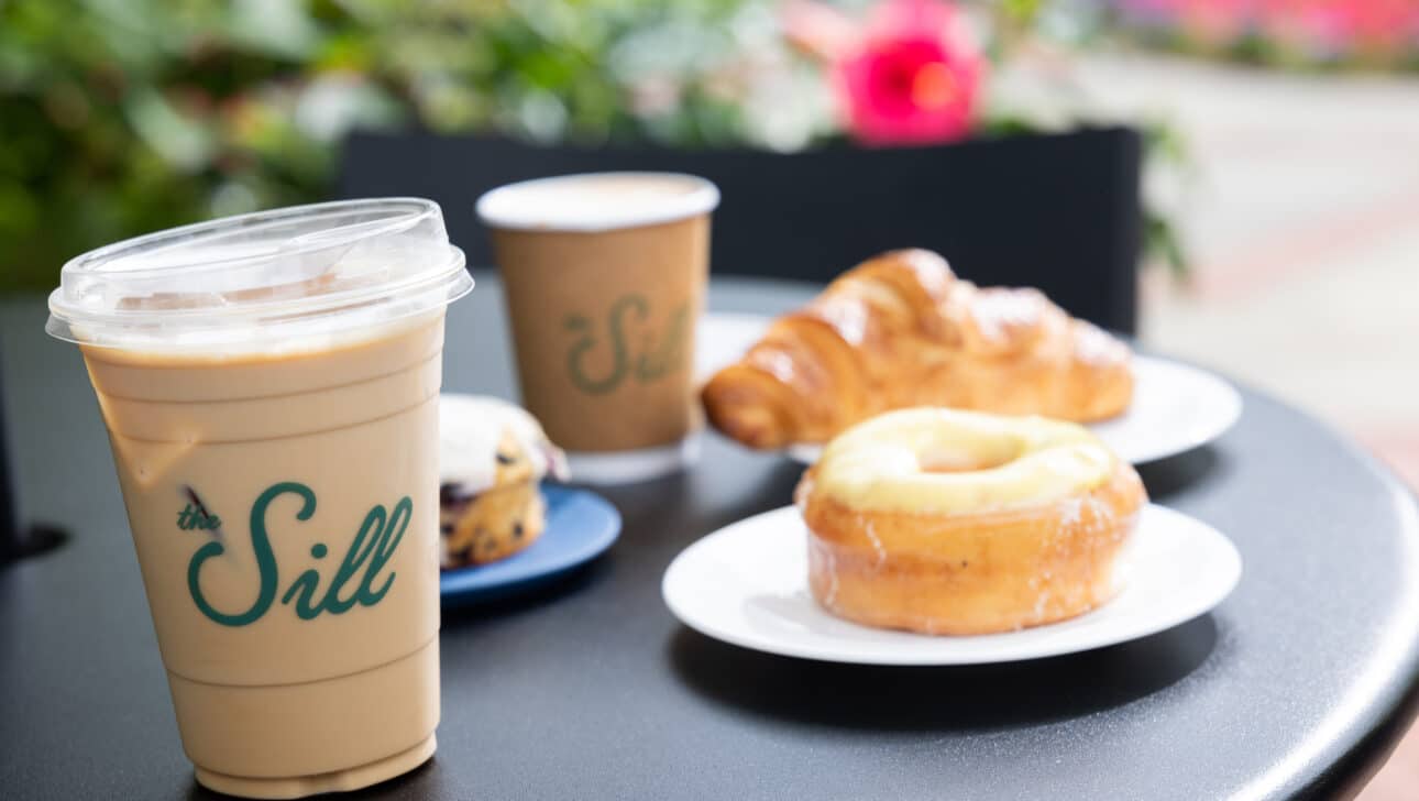 Iced coffee, hot coffee, a scone, a croissant, and a glazed donut on a black outdoor table with greenery and flowers in the background.