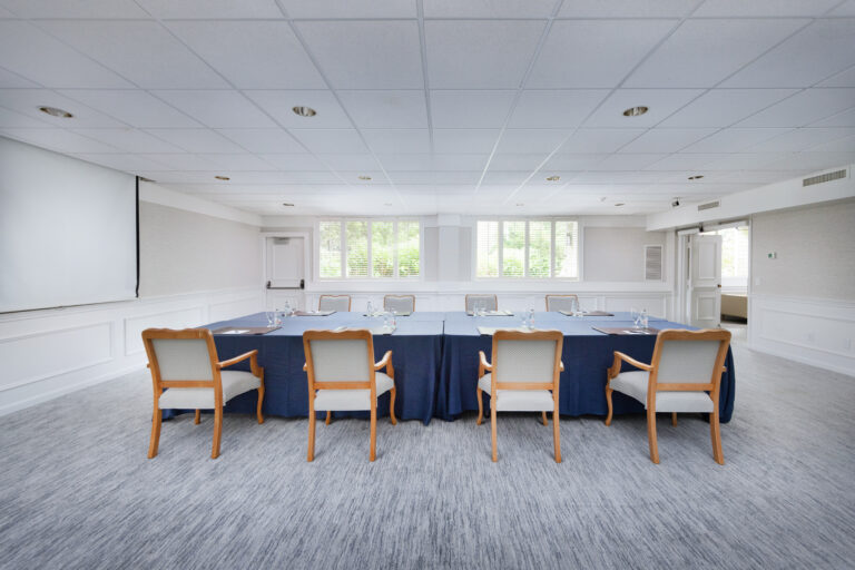 A versatile conference room with six wooden chairs around a navy-covered rectangular table, perfect for meetings or events, facing a projection screen and large windows.