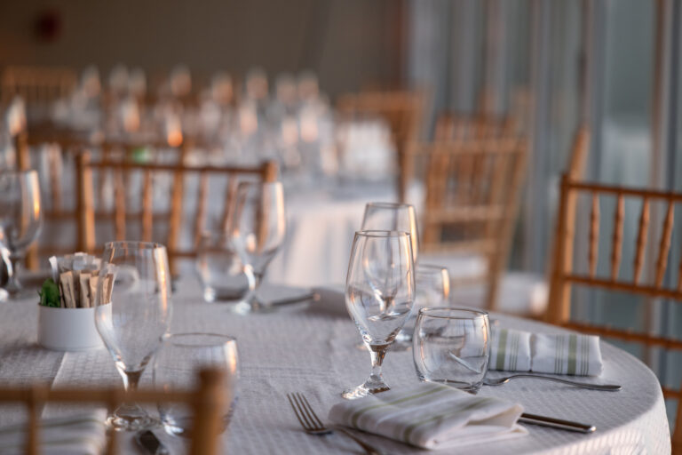 Round tables set with white tablecloths, folded napkins, glassware, and cutlery, surrounded by wooden chairs in a softly lit event space at Wequassett Resort.