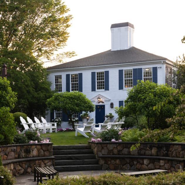 A large blue and white colonial-style house, reminiscent of a Cape Cod hotel, is surrounded by lush gardens, trees, and white Adirondack chairs arranged on the lawn.