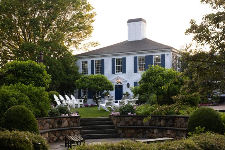 A large blue and white colonial-style house, reminiscent of a Cape Cod hotel, is surrounded by lush gardens, trees, and white Adirondack chairs arranged on the lawn.
