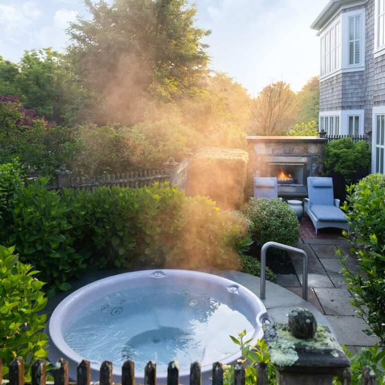 A steaming hot tub sits in a garden patio area beside a house, surrounded by greenery, with lounge chairs and an outdoor fireplace—evoking the luxurious ambiance of a Cape Cod resort.