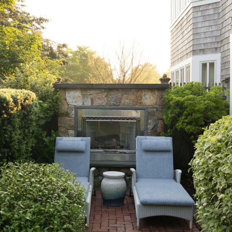 Two cushioned lounge chairs with a small round table between them are set on a brick patio in front of an outdoor stone fireplace, providing a relaxing retreat near the renowned Cape Cod golf course and 28 Atlantic restaurant.